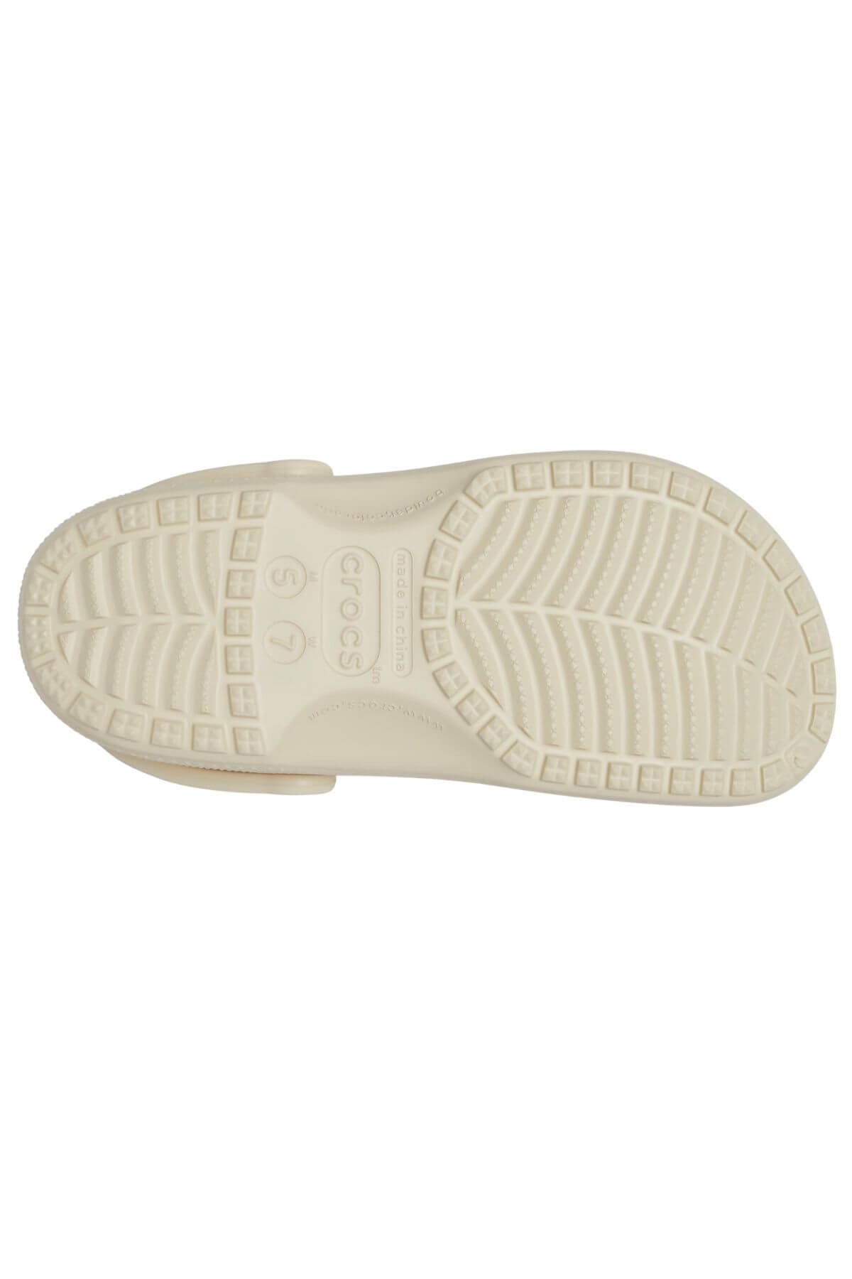 The image displays the sole of a light-colored Crocs Classic Clog sandal, featuring a textured pattern with small rectangular grips and a zigzag design for secure footing. It also includes size markings and the brand name.