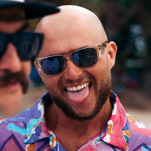 A bald man with a beard and SafeStyle Fusions Champagne Frame Polarised Lens sunglasses playfully sticks out his tongue. His colorful, patterned shirt adds to the lively scene. Another person in sunglasses is partially visible beside him, but the focus remains on him in the foreground.