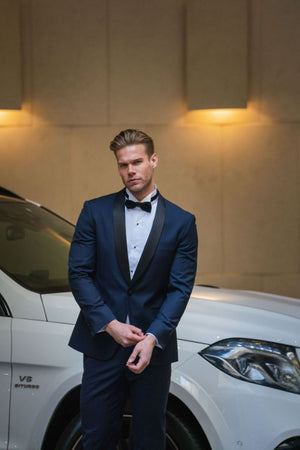 A man in a navy tuxedo and a Daniel Hechter Wing Stud Dinner Shirt stands in front of a white car with a serious expression. The car is parked indoors, and warm lighting illuminates the background, capturing the essence of formal occasions.