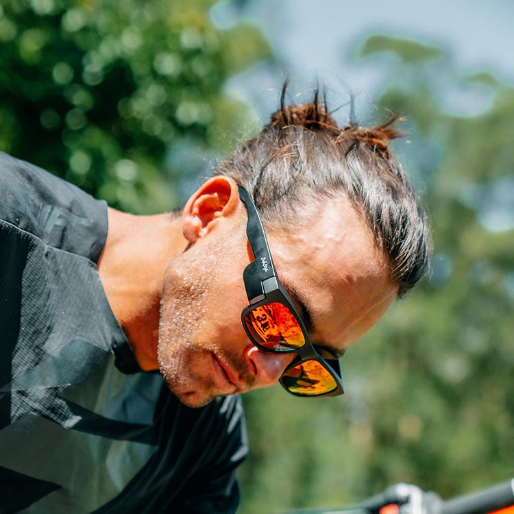 A person with long hair tied in a bun is seen outdoors sporting SafeStyle Fusions Matte Black Frame Mirror Red Polarised Lens sunglasses and a dark shirt. The background highlights trees and greenery under a clear sky.
