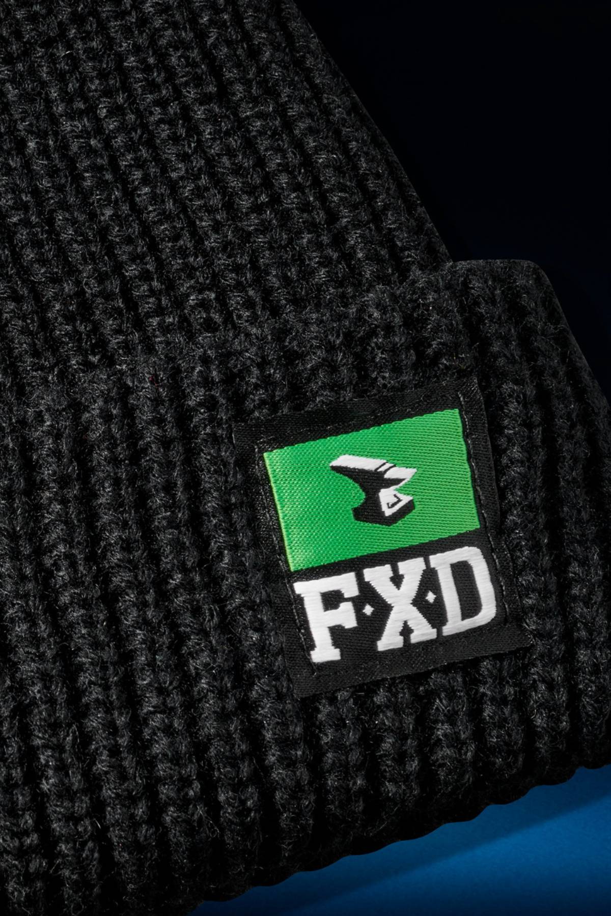 Close-up of the FXD CP-10 Knit Ribbed Beanie in black, made from 100% acrylic, with a rectangular patch featuring a green and black stylized bird logo and bold white "FXD" lettering.