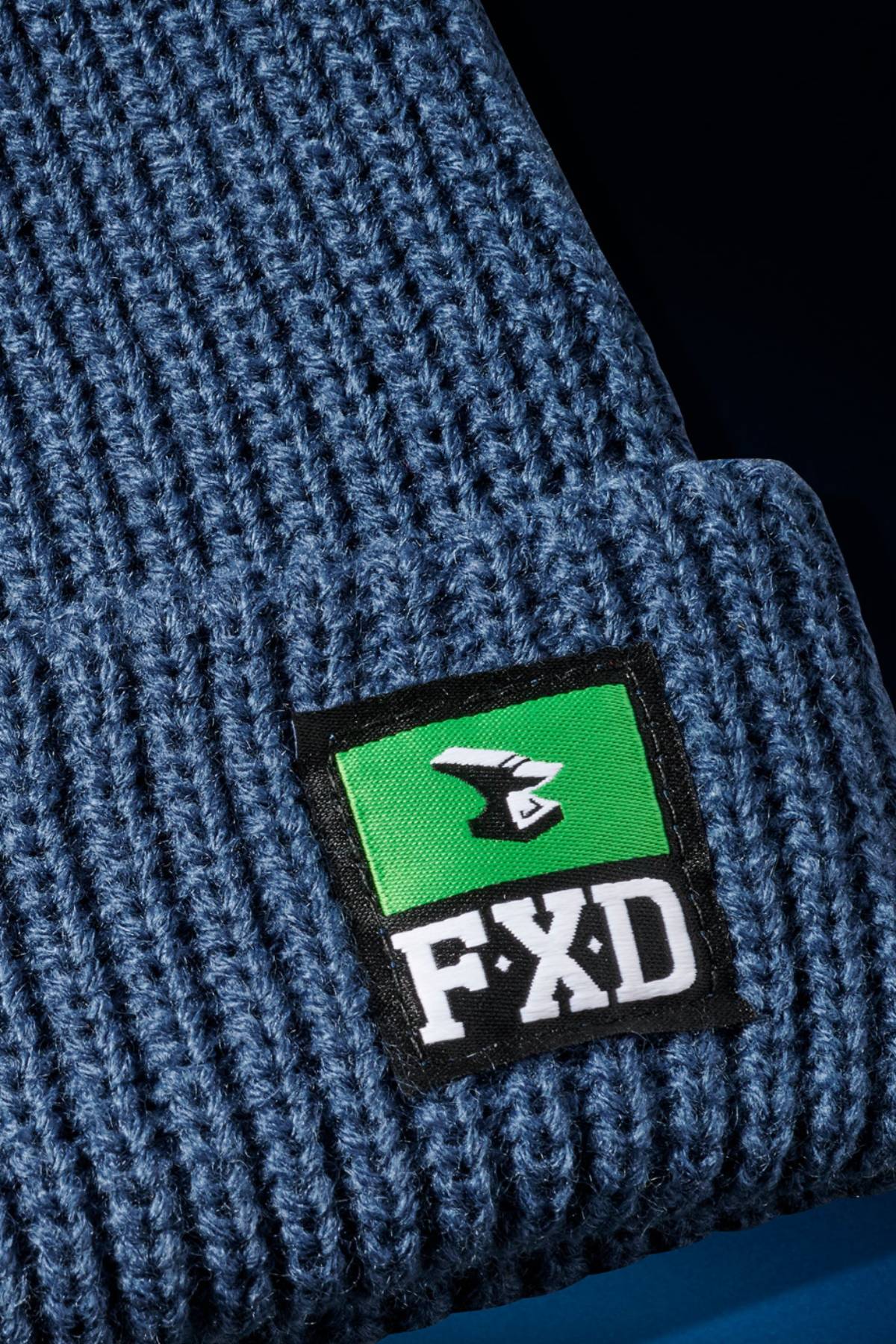 Close-up of the FXD CP-10 Knit Ribbed Beanie in blue with a black, green, and white FXD patch featuring a stylized bird logo on the turned-up brim and durable ribbed knit construction.