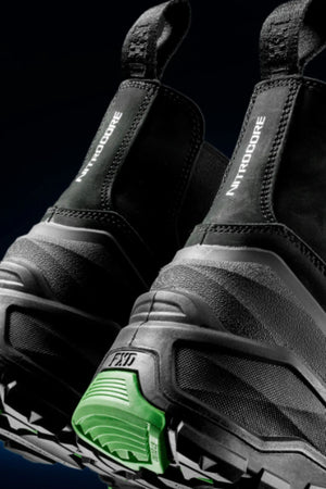Close-up of the back of two black FXD DURA X-3 Slip On Work Boots with thick, rugged soles. "NITROCORE" appears on the heel tab and one sole displays a green accent near the heel.