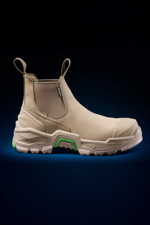 The FXD DURA X-3 Slip On Work Boots feature a beige design with pull tabs, rugged tan NITROCORE sole for added grip, and green accents, shown against a dark gradient backdrop.