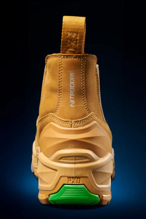 The FXD DURA X-3 Slip On Work Boots feature a tan industrial design, "NITROCORE" text on the back, chunky textured sole, and a green heel accent for style and puncture-resistant protection, shown against a dark gradient background.