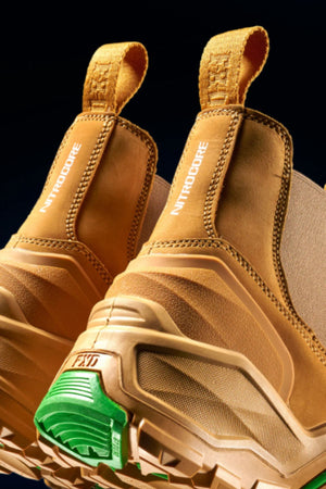 Close-up of FXD DURA X-3 Slip On Work Boots in tan, featuring rugged, puncture-resistant soles with green and white tread details and "NITRODORE" on the side pull-loops, set against a dark background.