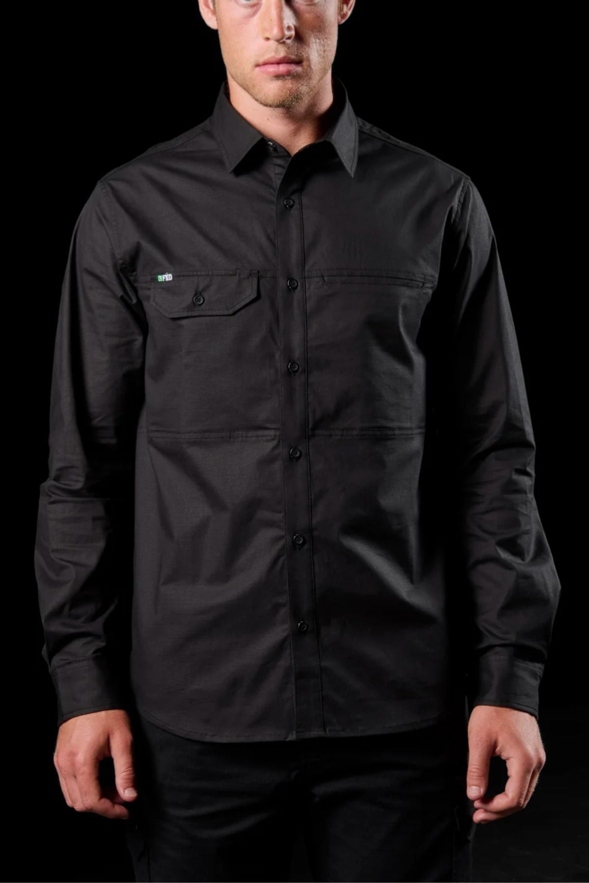 A man stands against a black background wearing the FXD LSH-3 Vented Work Shirt—a black, long-sleeve, button-up shirt with a left chest pocket. Only his upper body and face are visible.
