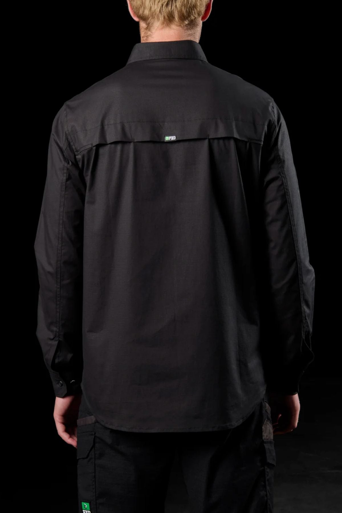 A blond-haired person is seen from behind in a solid black setting, wearing the FXD LSH-3 Vented Work Shirt and black pants with green and white logo tags. The outfit features lightweight stretch fabric for all-day comfort.
