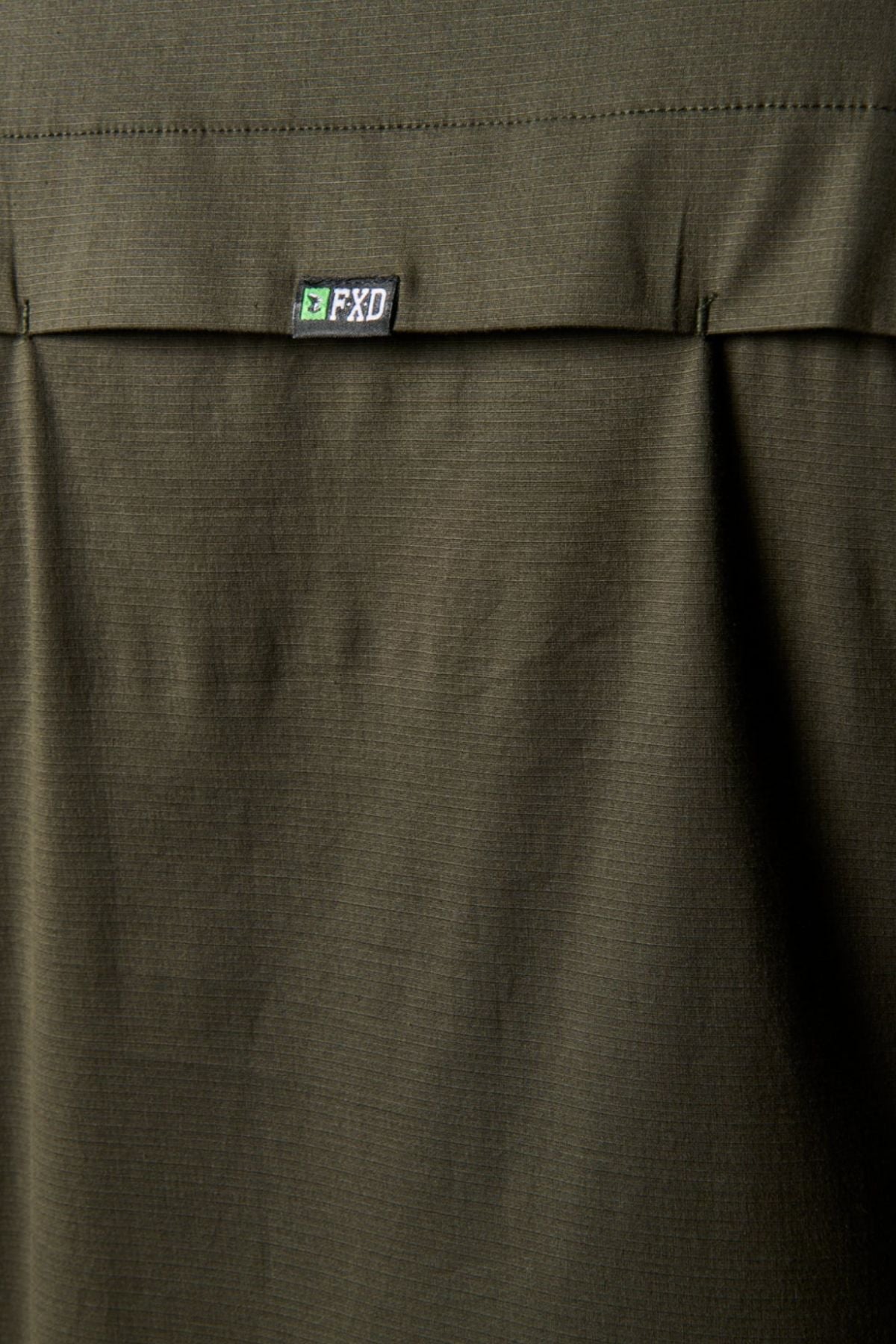 Close-up of olive green stretch fabric with a flap and small black and white FXD logo tag, part of a pocket on the FXD LSH-3 Vented Work Shirt.