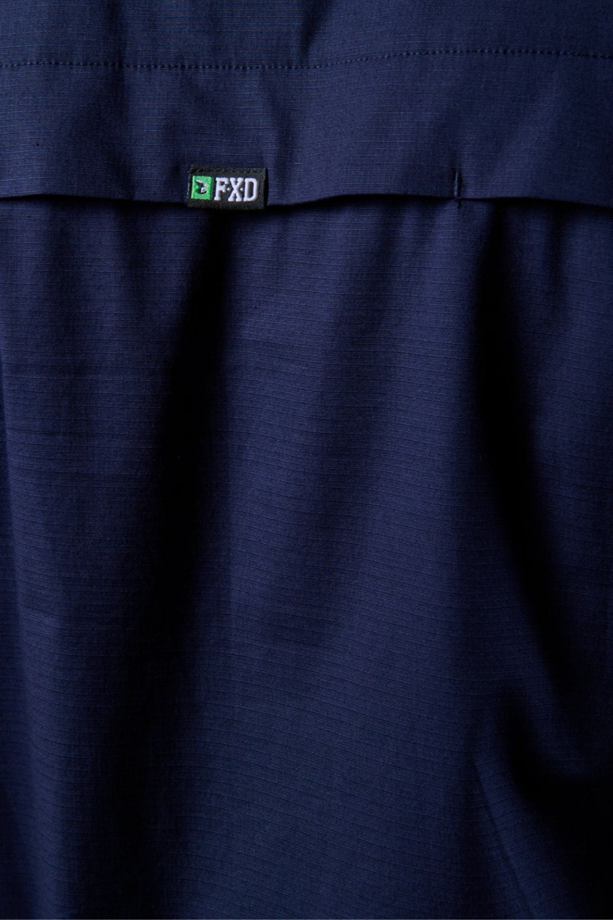 A close-up of the dark blue FXD LSH-3 Vented Work Shirt's lightweight stretch fabric, featuring a small black, green, and white FXD brand tag sewn near the top edge.