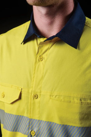 A close-up of a person wearing the FXD LSH-4T Work Shirt, showcasing its yellow button-up style, dark blue collar, and segmented reflective tape on the chest, against a dark background.