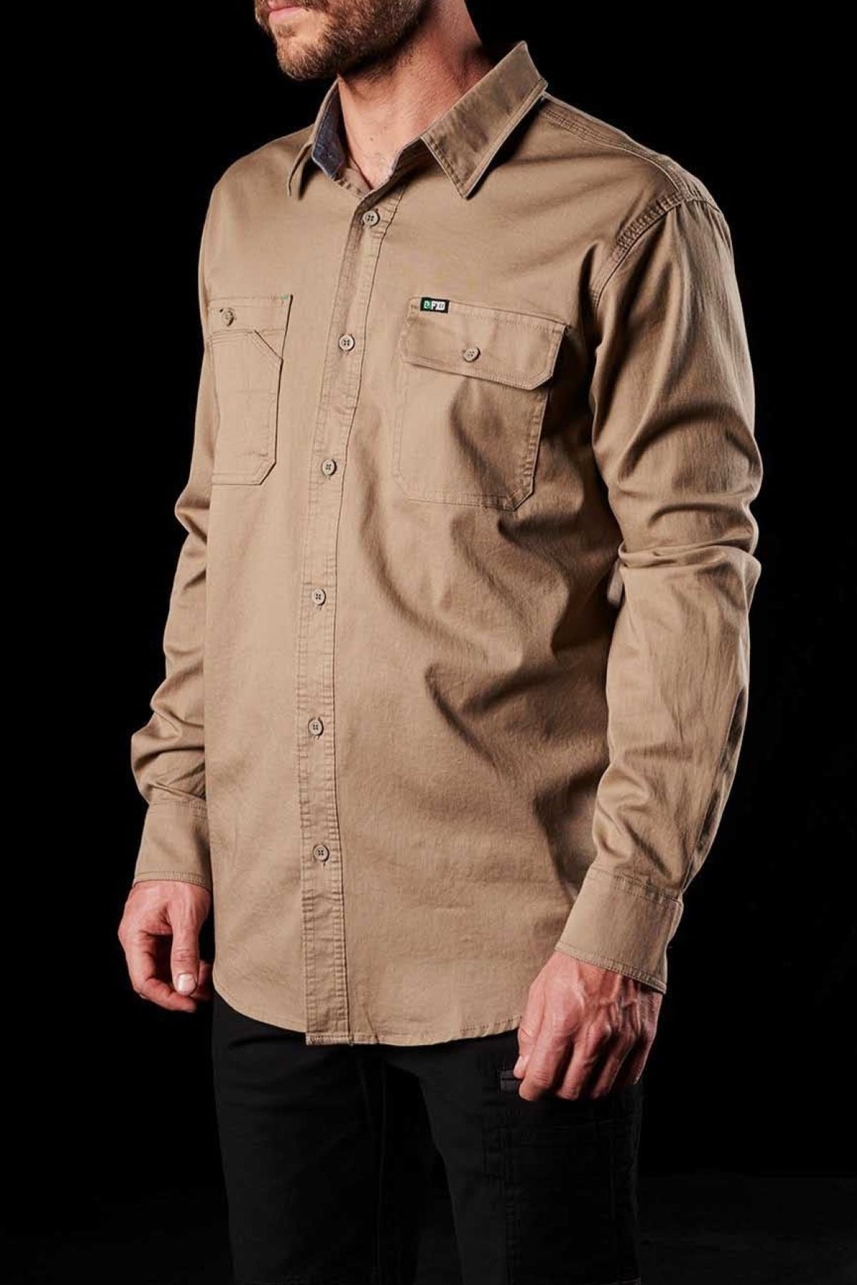 A person is wearing a comfortable FXD LSH-1 Stretch Work Shirt with durable triple needle seams, featuring a long-sleeved beige button-up design and two chest pockets with button flaps. The shirt is paired with dark pants, set against a solid black background.