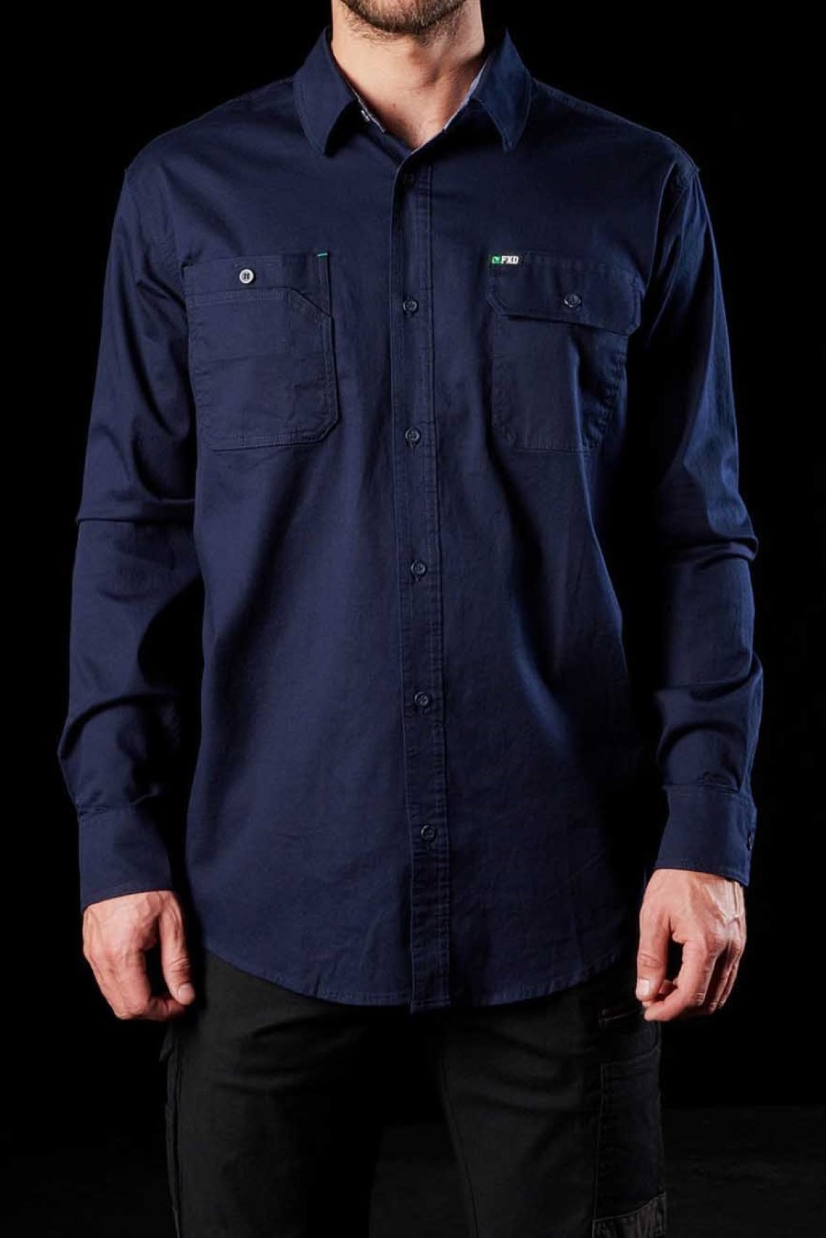 A person wearing the FXD LSH-1 Stretch Work Shirt in navy blue, characterized by its two front chest pockets with buttoned flaps that highlight its durability, stands against a black background. The shirt's long sleeves and practical design are visible with the person's hands relaxed at their sides. Only the torso and arms are shown.