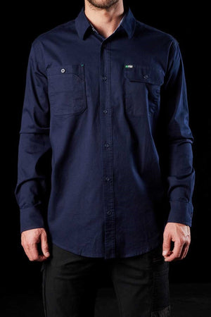 A person wearing the FXD LSH-1 Stretch Work Shirt in navy blue, characterized by its two front chest pockets with buttoned flaps that highlight its durability, stands against a black background. The shirt's long sleeves and practical design are visible with the person's hands relaxed at their sides. Only the torso and arms are shown.