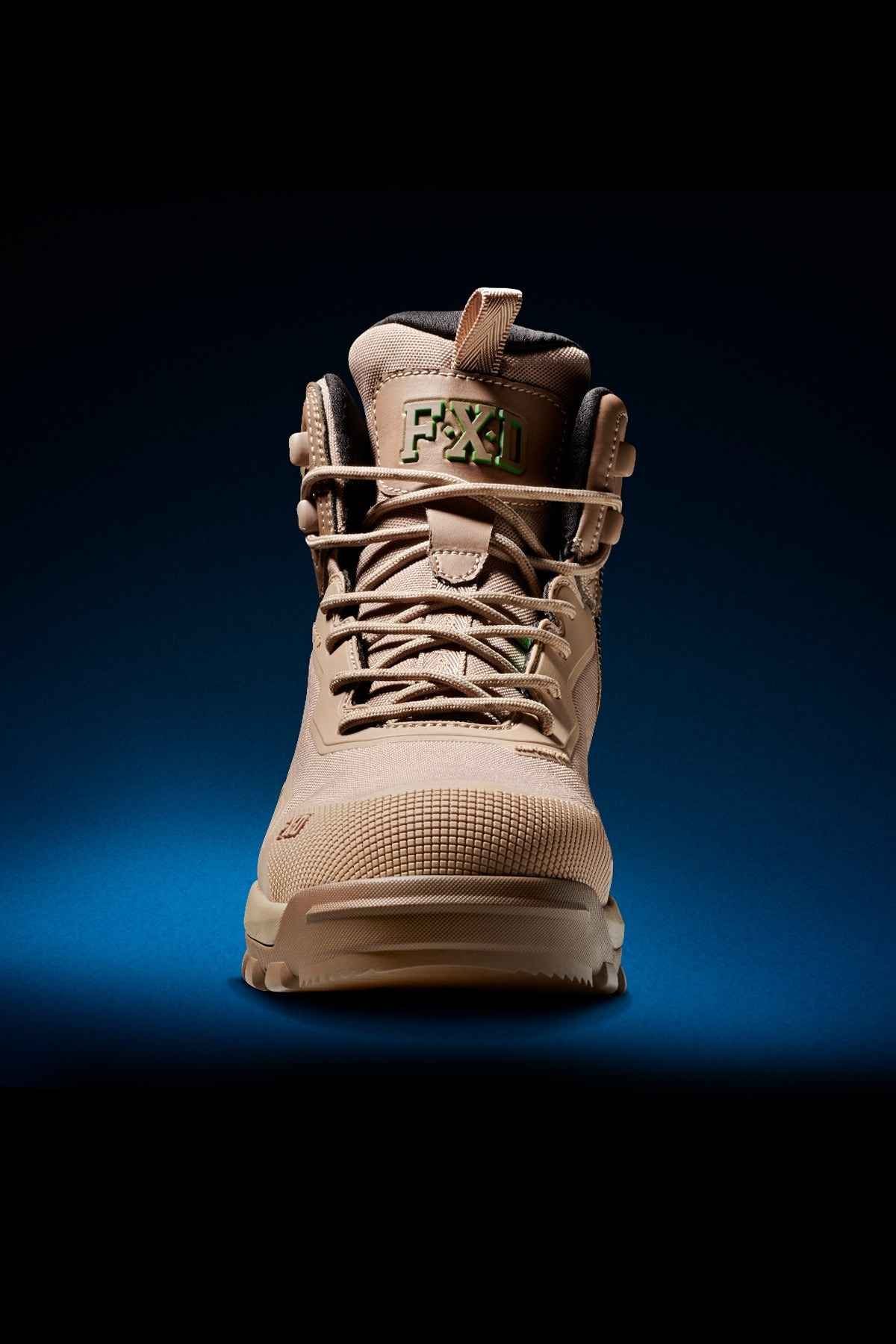 A close-up view of the tan-colored FXD WB-6 Work Boot showcases its detailed laces, distinctive green FX1 logo on the tongue, and rugged sole. The boot is designed with a PORON heel insert for added comfort and meets safety standards. It is well-lit against a dark gradient background, accentuating its texture and design features.