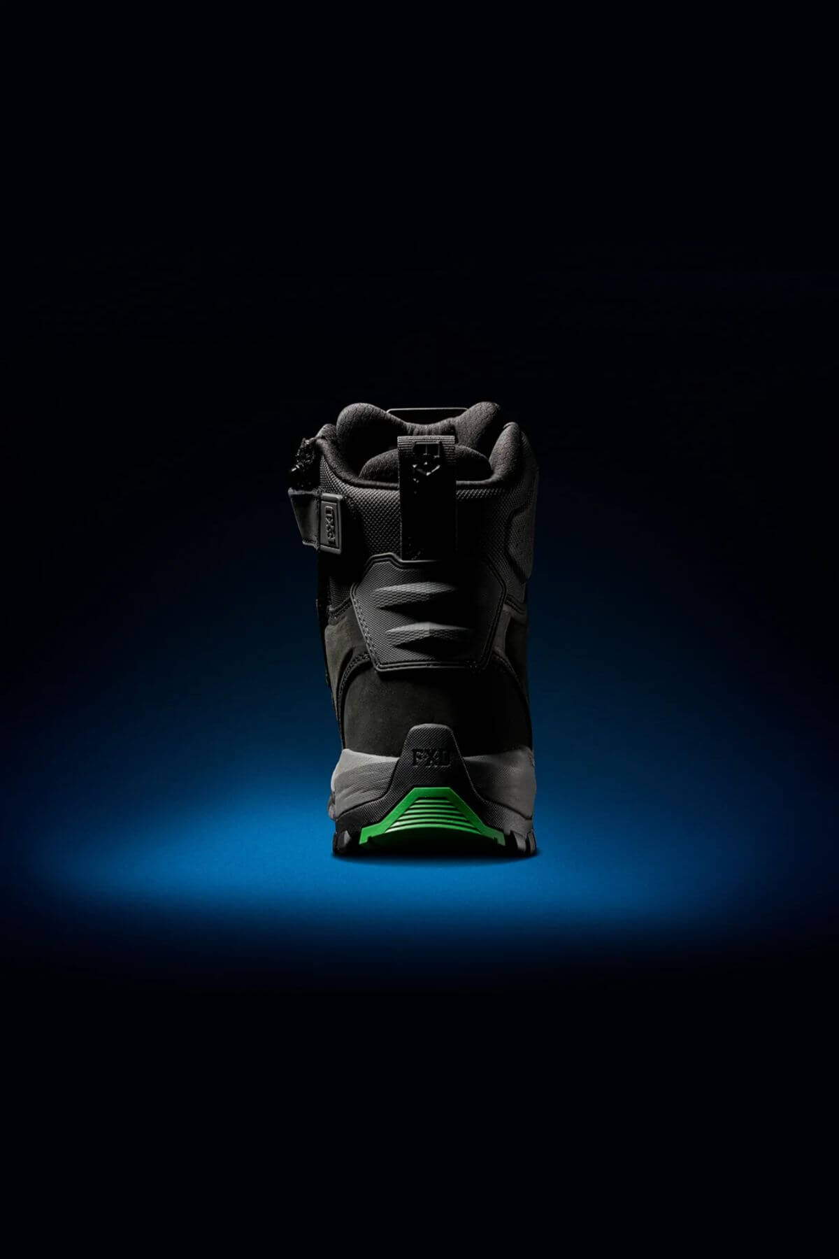 A single FXD WB-1 Work Boot in black is showcased against a dark background with a blue spotlight effect beneath it. The heel of the boot is prominently displayed, featuring a green sole and intricate design details on the upper, emphasizing its slip-resistant feature.