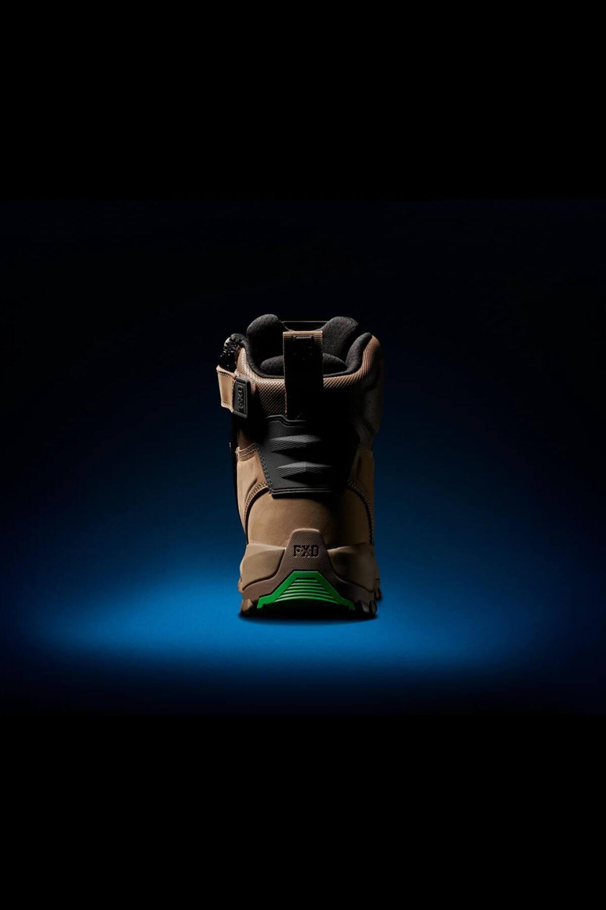 The FXD WB-1 Work Boot is showcased from the back, featuring a tan and black color scheme with a green sole. Set against a dark background illuminated by a blue spotlight, this boot highlights its rugged design with sturdy construction, a safety toe, and a buckle strap.
