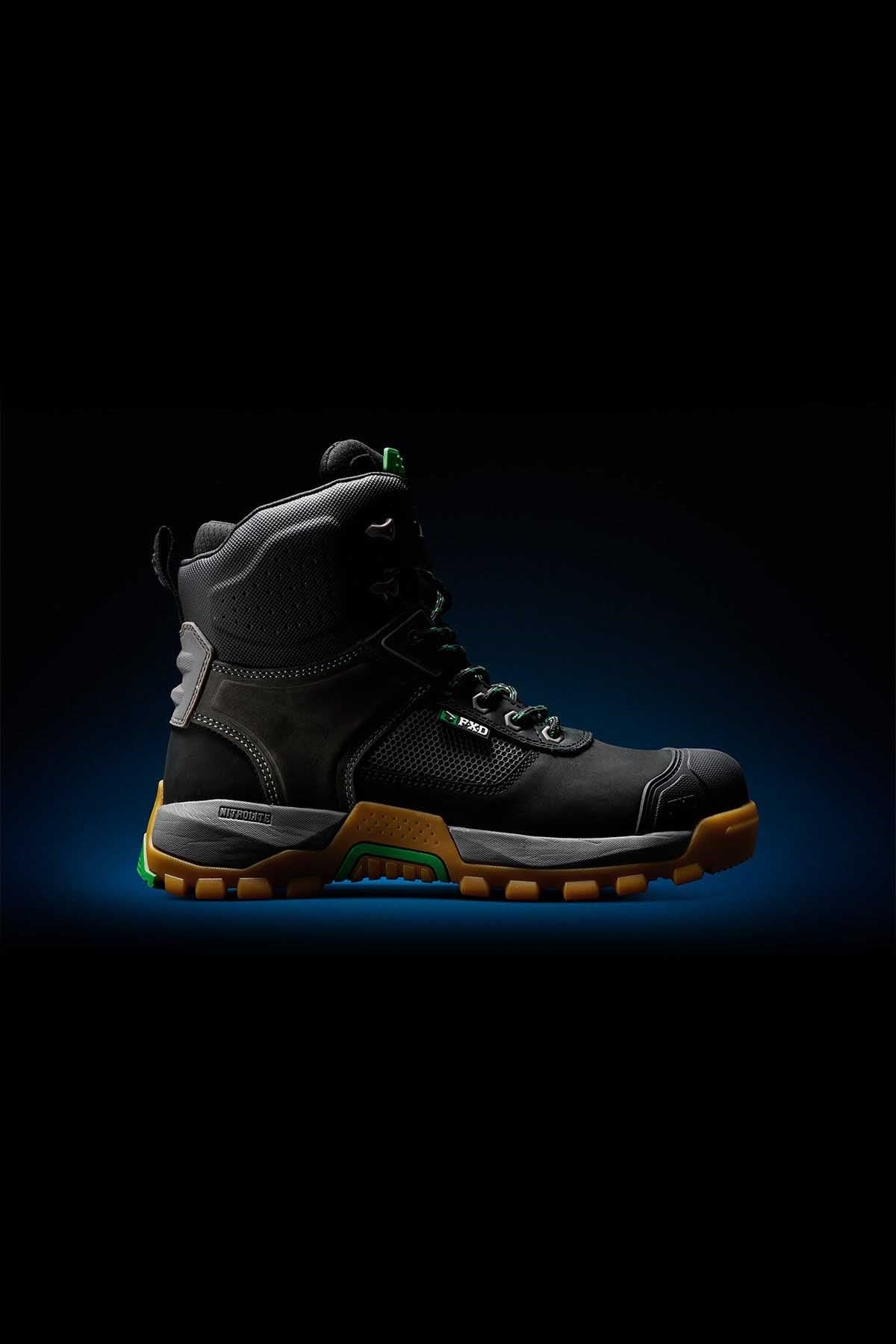 The FXD WB-1 Work Boot, a black high-top industrial work boot with green and brown accents, is displayed on a dark gradient background. Featuring NITROLITE midsole cushioning, the boot boasts a rugged sole, composite safety toe, and metal eyelets for laces, suggesting durability and protection.