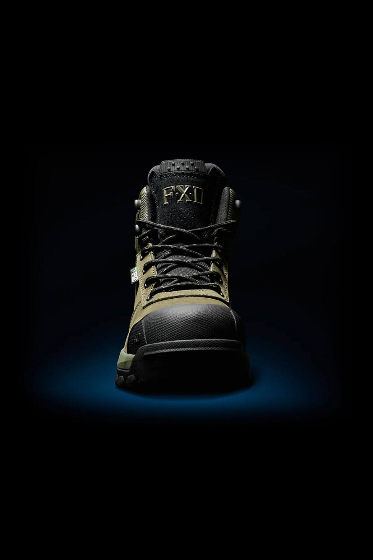 An FXD WB-2 Work Boot in black and olive green, with a composite safety toe and black laces, is centered under a spotlight on a dark background. The "FXD" logo appears on the tongue.