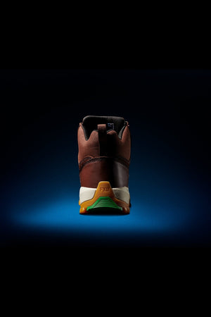 The FXD WB-3 Work Boot is a brown sneaker-style work boot with Premium Leather, a white and yellow sole, green tread on the heel, and "FXD" on the heel tab, shown from the back against a dark blue gradient background.