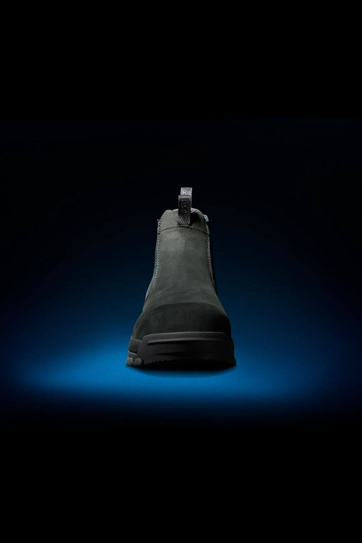The FXD WB-4 Work Boot in dark green, featuring a safety toe and a pull-tab at the back, is showcased under dramatic studio lighting against a black background with a subtle blue glow beneath it. The Nubuck leather boot's rugged sole and front profile are visible, highlighting its sturdy design.