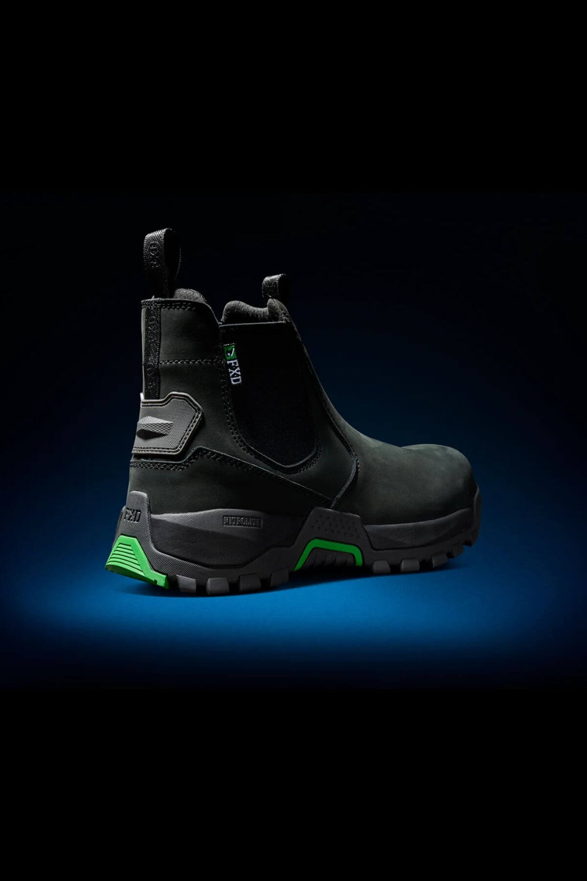 The FXD WB-4 Work Boot, featuring a black Nubuck leather construction with a striking green accent on the heel and sole, is set against a dark gradient background. This work boot includes a pull tab, side elastic panels, and a rugged, durable sole equipped with a safety toe, designed for heavy-duty or industrial use.