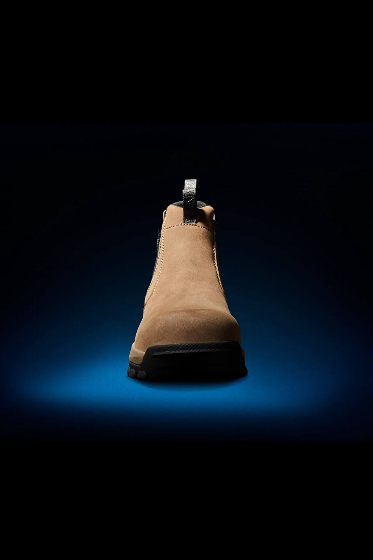 A single FXD WB-4 Work Boot in tan color, featuring slip-resistant soles, is showcased against a dark backdrop with a blue spotlight beneath it, emphasizing the shoe. The boot includes a pull tab at the back and exhibits stitching details around the ankle area.