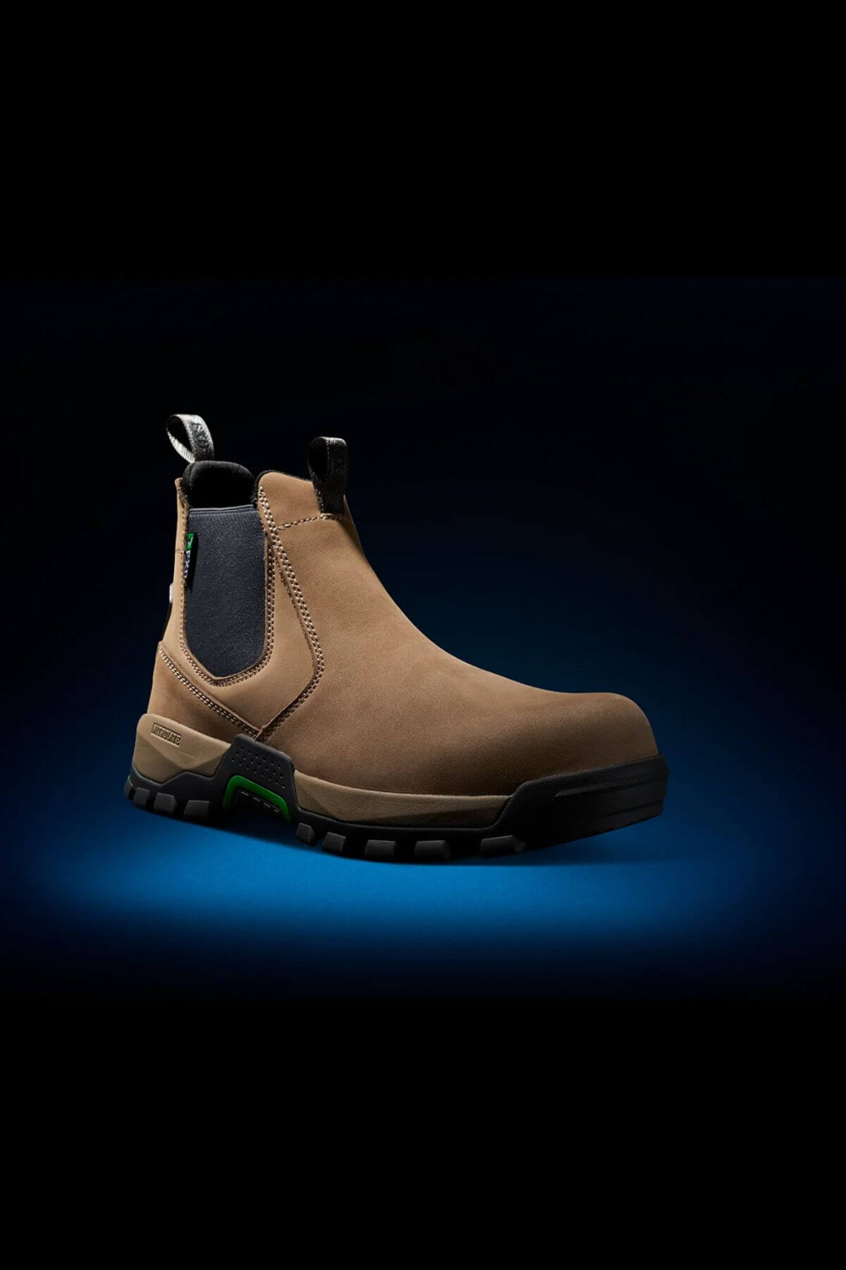 The FXD WB-4 Work Boot is showcased against a dark gradient background highlighted by a blue spotlight underneath. This tan-colored, slip-on work boot features a black pull tab, elastic panels at the ankle, and a durable black rubber sole. The safety toe offers additional protection, while green accents on the sides add a stylish touch.