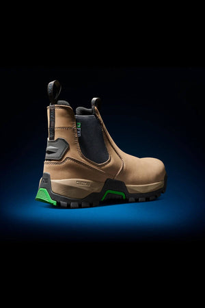 Presenting the FXD WB-4 Work Boot: a brown slip-on work boot showcasing a rugged design with a durable black and green slip-resistant sole. The boot features black accents around the elastic sides and heel, a reinforced safety toe for extra protection, and a convenient pull tab at the back. The dark background accentuates the boot, which stands out on an illuminated blue surface.