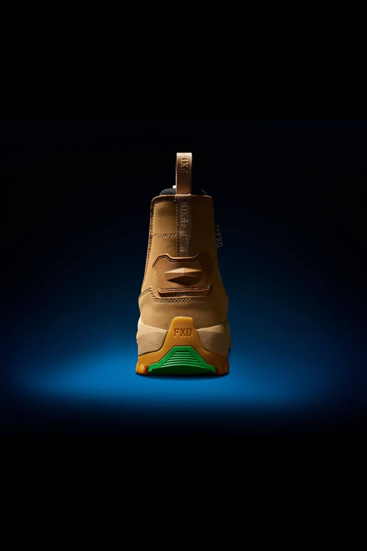The FXD WB-4 Work Boot, a single beige work boot with a green sole, is displayed against a dark gradient background. The heel of the boot, featuring a safety toe, faces the viewer and highlights a pull tab at the back. The sole and part of the upper showcase subtle design details.