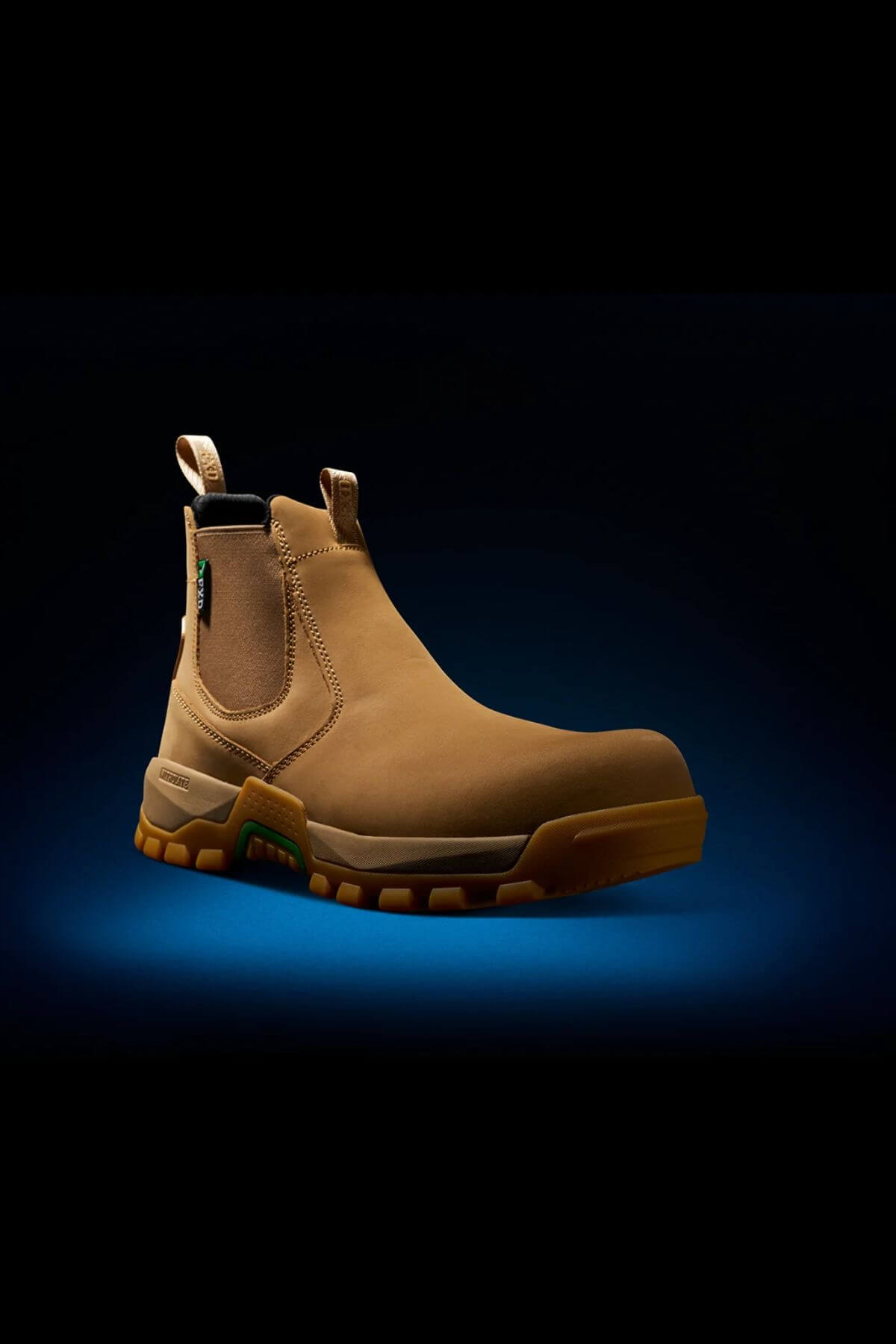 The FXD WB-4 Work Boot is a tan-colored, slip-on work boot that features a safety toe and convenient pull tabs on the front and back. It boasts rugged rubber soles, elastic side panels, and is displayed against a dark background with blue lighting highlighting it from beneath.