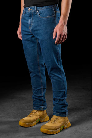 A person wearing FXD WD-4 durable straight fit blue jeans made with COOLMAX EcoMade fabric, a black shirt, and tan sneakers stands against a dark background. The image is cropped from the shoulders down.