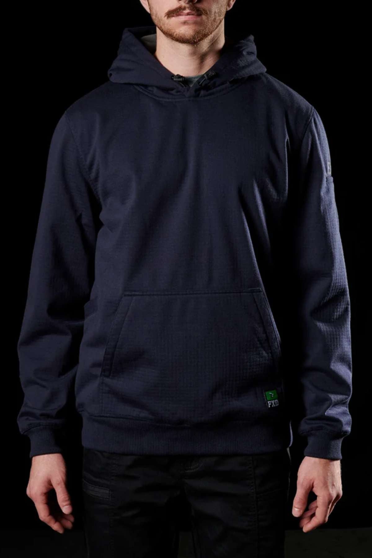 A man is standing against a black background wearing a dark navy FXD WF-1 Hoodie with a front pocket and black pants. The hoodie, made from windproof fleece, features a small green embroidered patch near the pocket. The man's face is partially visible, highlighting the clothing.