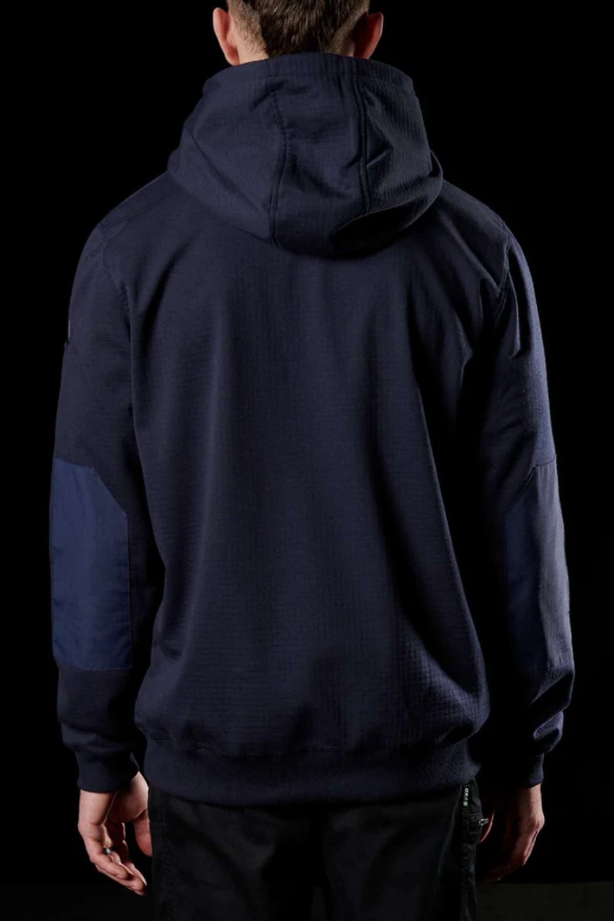 A person is seen from the back, clad in a navy blue FXD WF-1 Hoodie with the hood up, against a black background. The windproof fleece hoodie showcases darker blue patches on the elbows. The person's arms hang down by their sides.