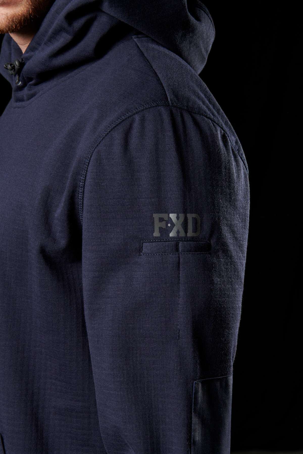 Close-up of a person wearing a dark navy FXD WF-1 Hoodie against a black background. The windproof fleece hoodie features the letters "FXD" on the upper sleeve and has visible seam detailing along the shoulder and arm. The hood is up, partially covering the head.