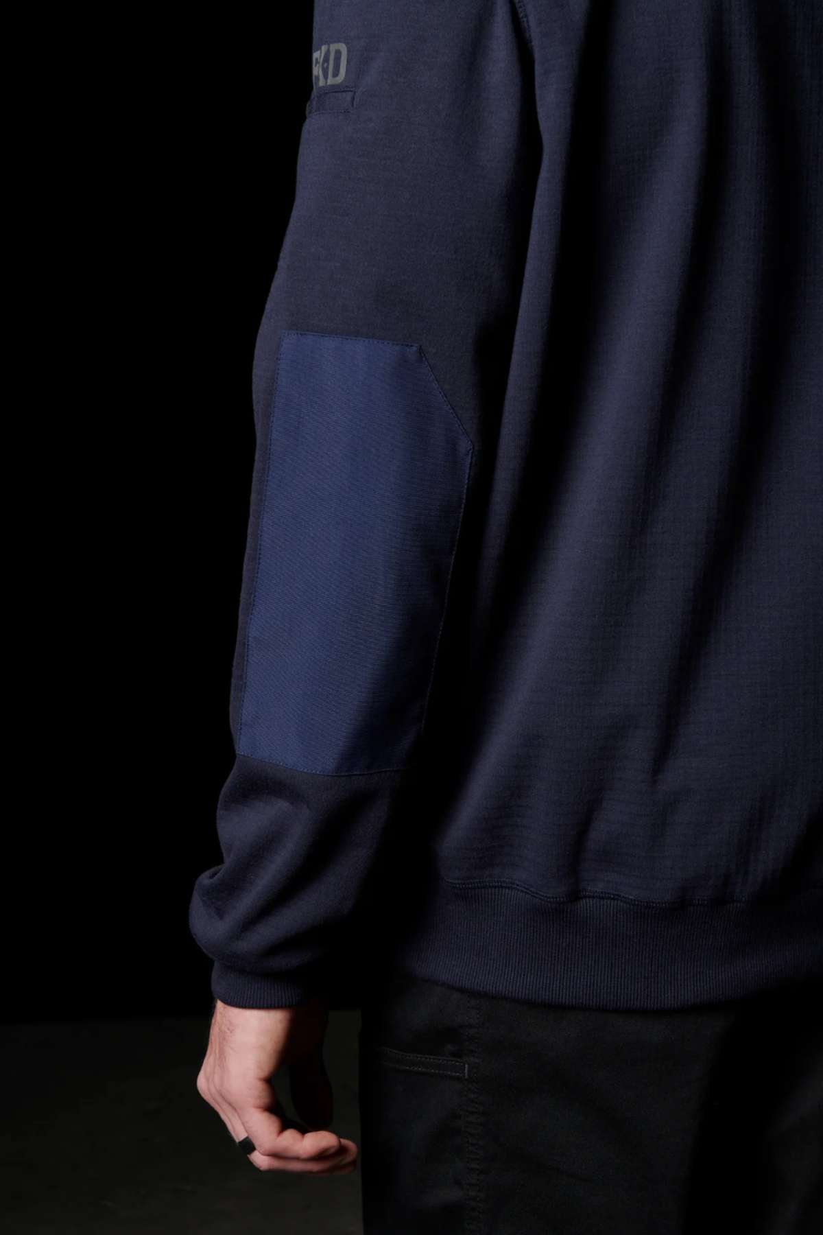 A close-up of a person wearing the FXD WF-1 Hoodie in dark navy blue, featuring a contrasting blue patch on the elbow. The person stands against a black background with just the back of the hoodie and part of their left arm visible, their hand resting casually by their side.
