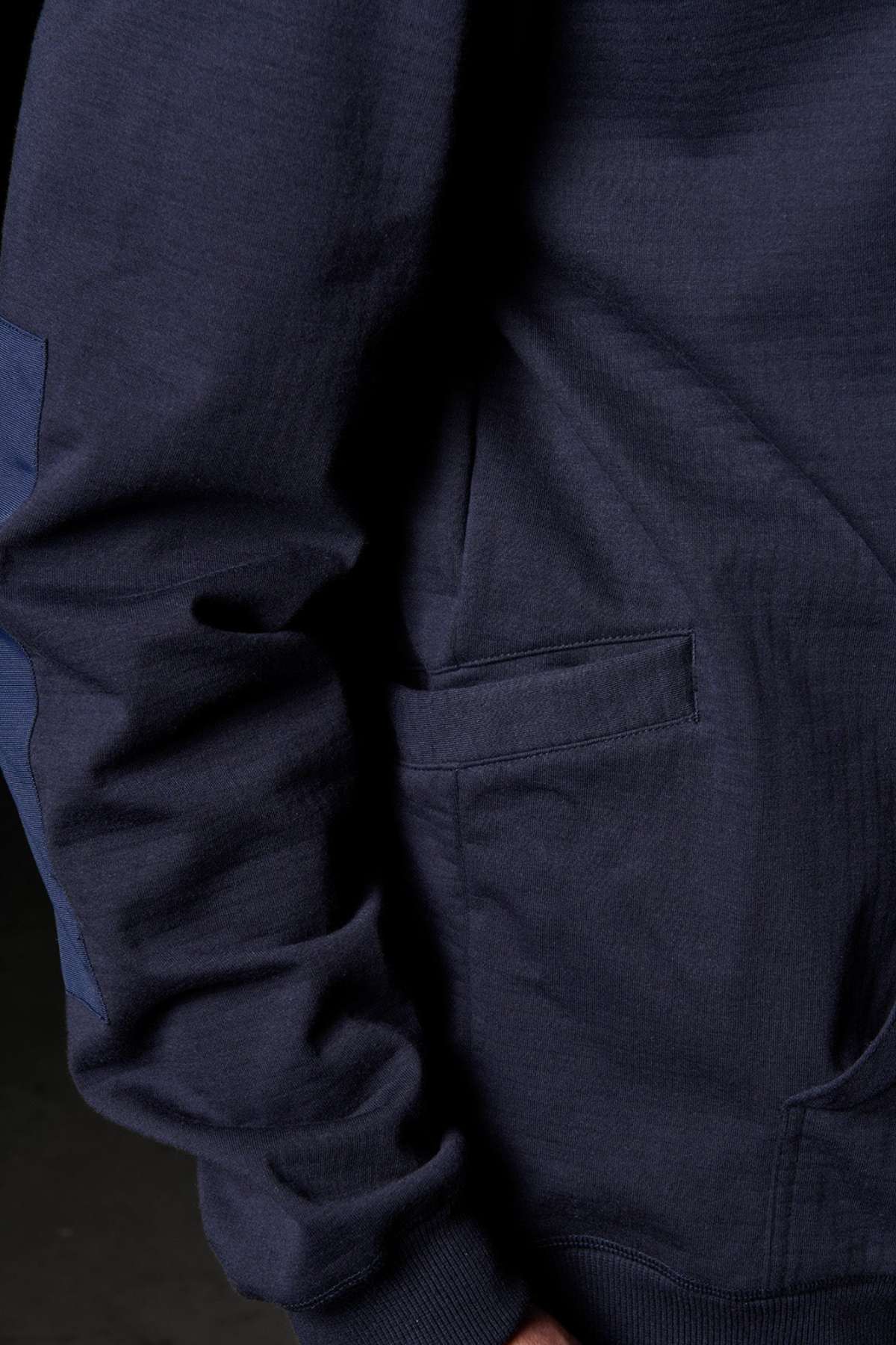 A close-up image of a person wearing an FXD WF-1 Hoodie in dark blue. One arm is visible, slightly bent at the elbow, and there is a side front pocket on the hoodie. The background is dark and out of focus.