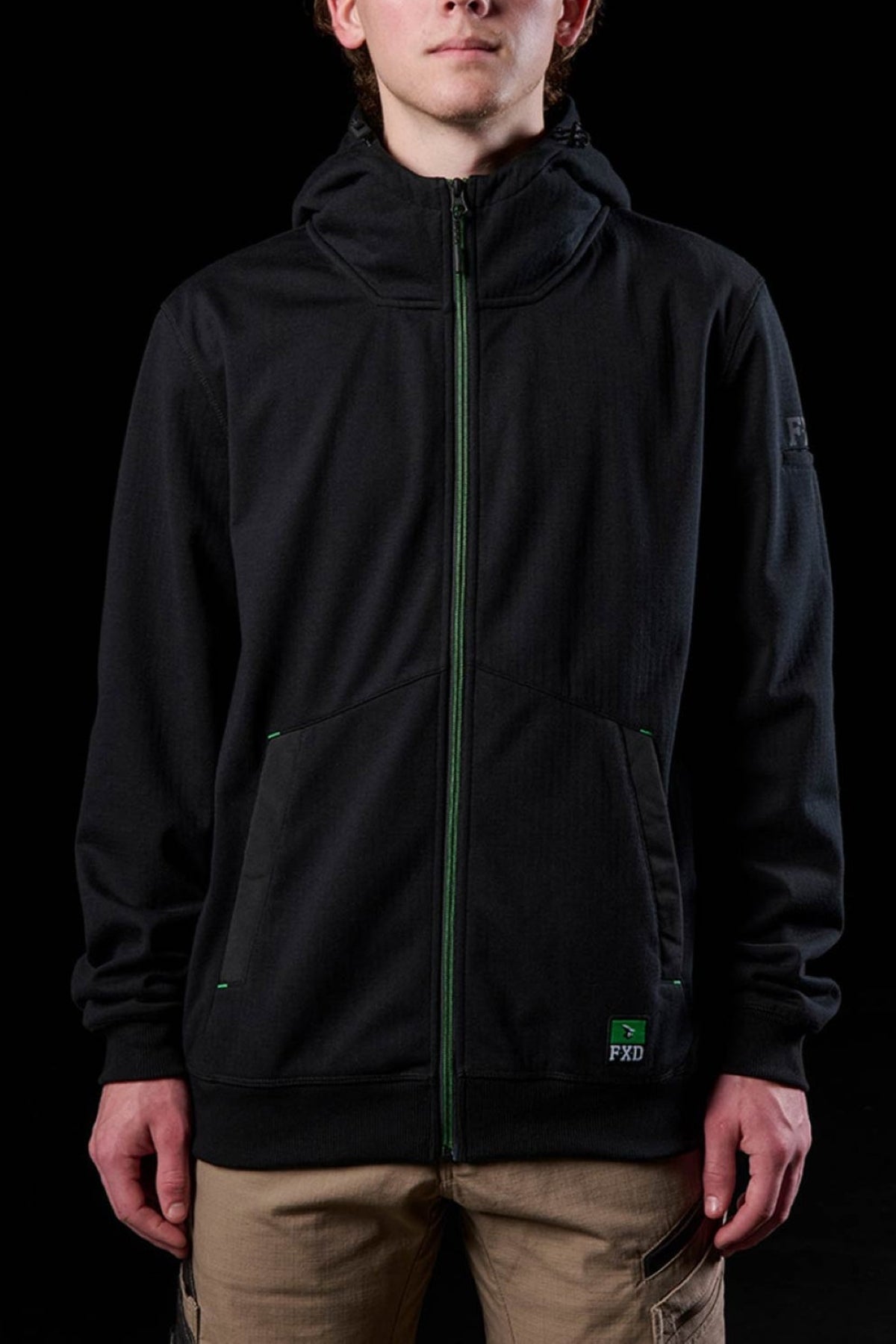 A person faces the camera against a black background, wearing the FXD WF-3 Jacket—a black water-resistant workwear hoodie with green accents and a small "FXD" logo by the pocket—paired with tan pants.