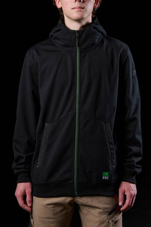 A person faces the camera against a black background, wearing the FXD WF-3 Jacket—a black water-resistant workwear hoodie with green accents and a small "FXD" logo by the pocket—paired with tan pants.
