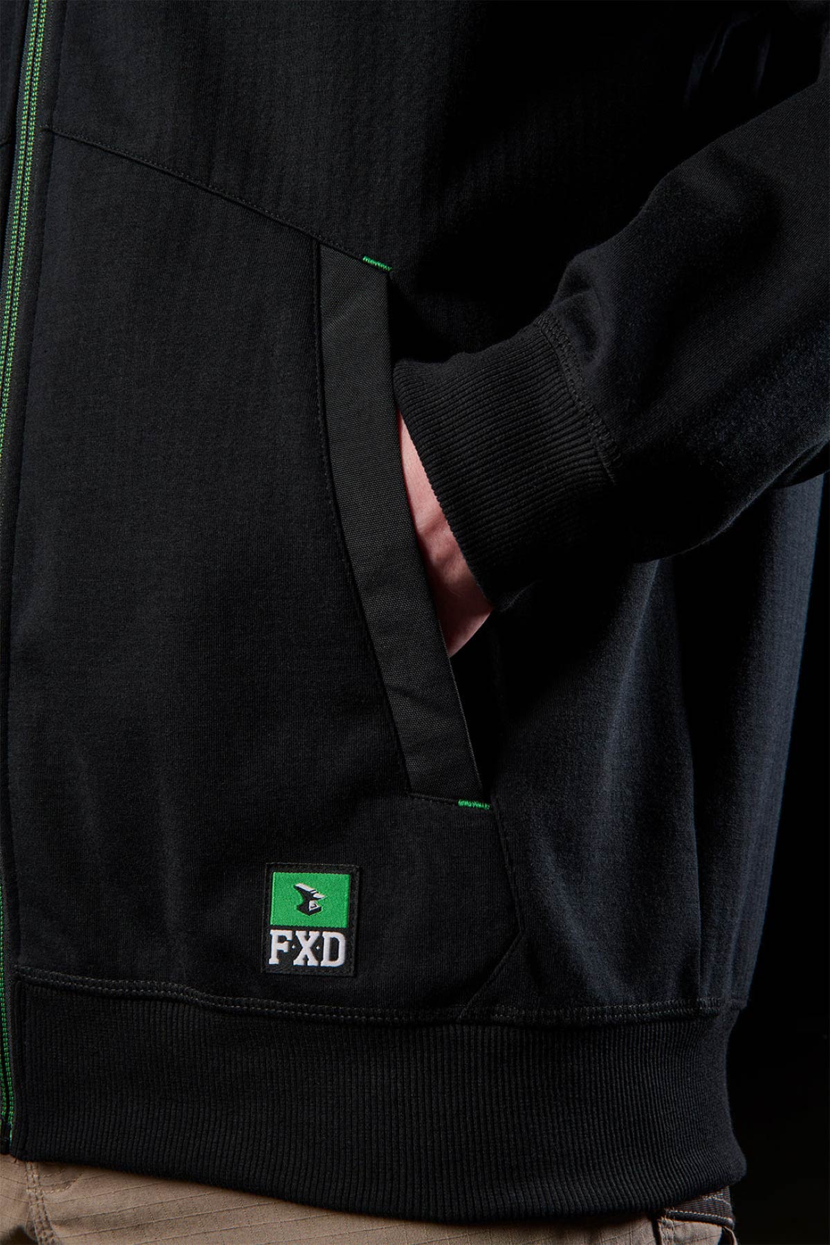 Wearing a black FXD WF-3 Jacket with green accents and the FXD logo near the hem, a person stands with their hand in the pocket. The water-resistant jacket is styled with tan workwear pants for a practical look.