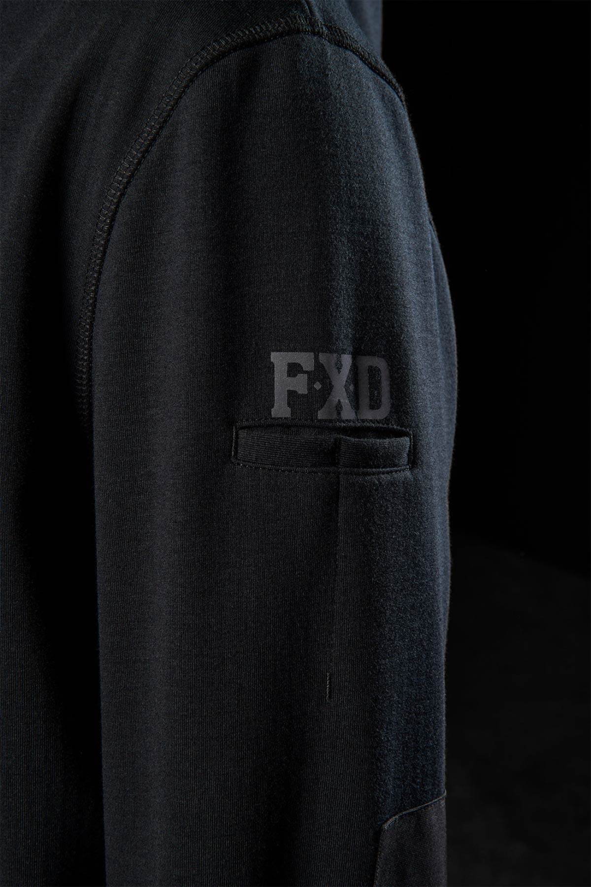 Close-up of a black FXD WF-3 Jacket sleeve shows a small pocket with the "FXD" logo above it. The dark background emphasizes the textured, high-quality design details characteristic of the FXD WF-3 Jacket.