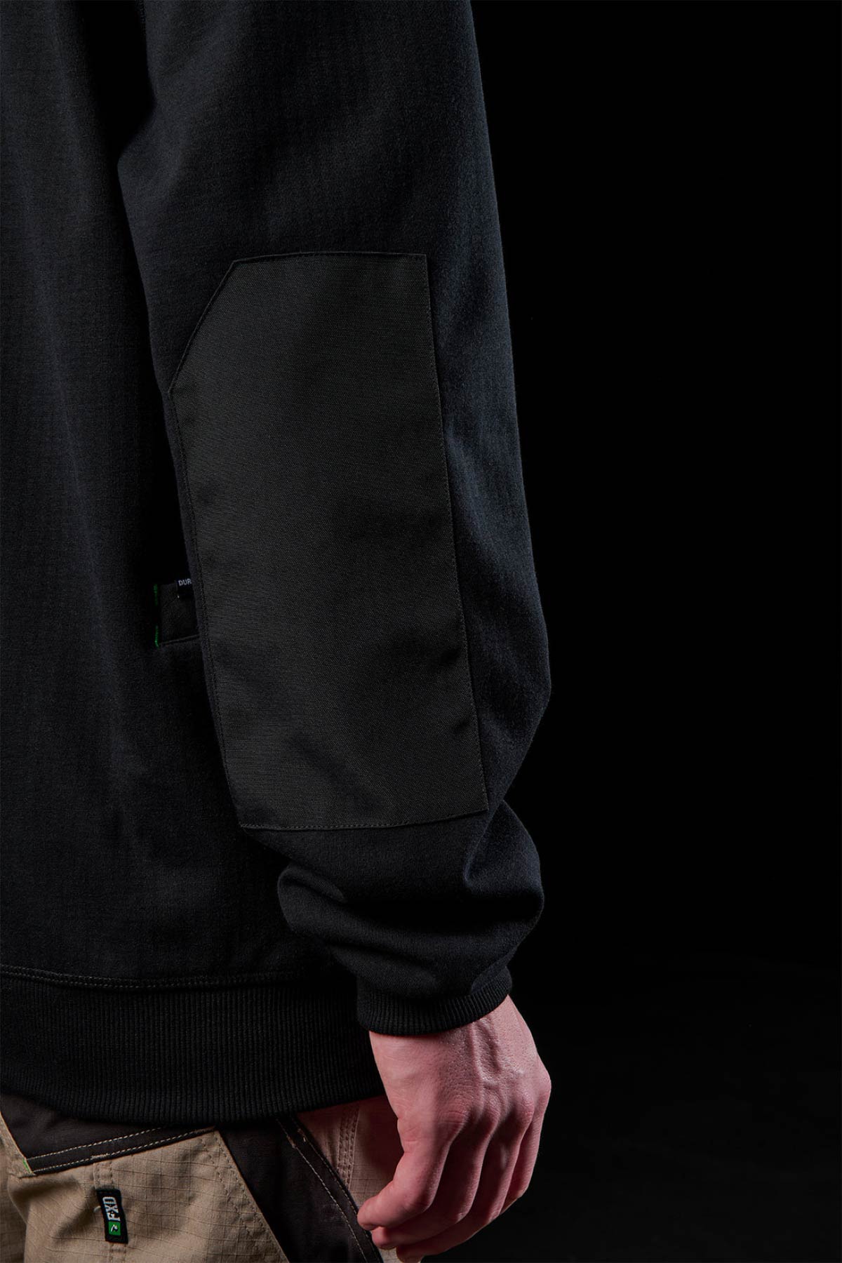 Close-up of a person in the FXD WF-3 Jacket with textured elbow patch and beige work pants, standing against a black background. Only their arm and hand are partially visible.