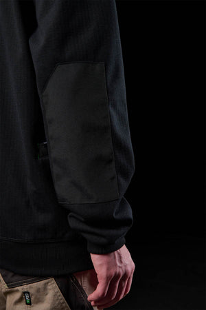 Close-up of a person in the FXD WF-3 Jacket with textured elbow patch and beige work pants, standing against a black background. Only their arm and hand are partially visible.