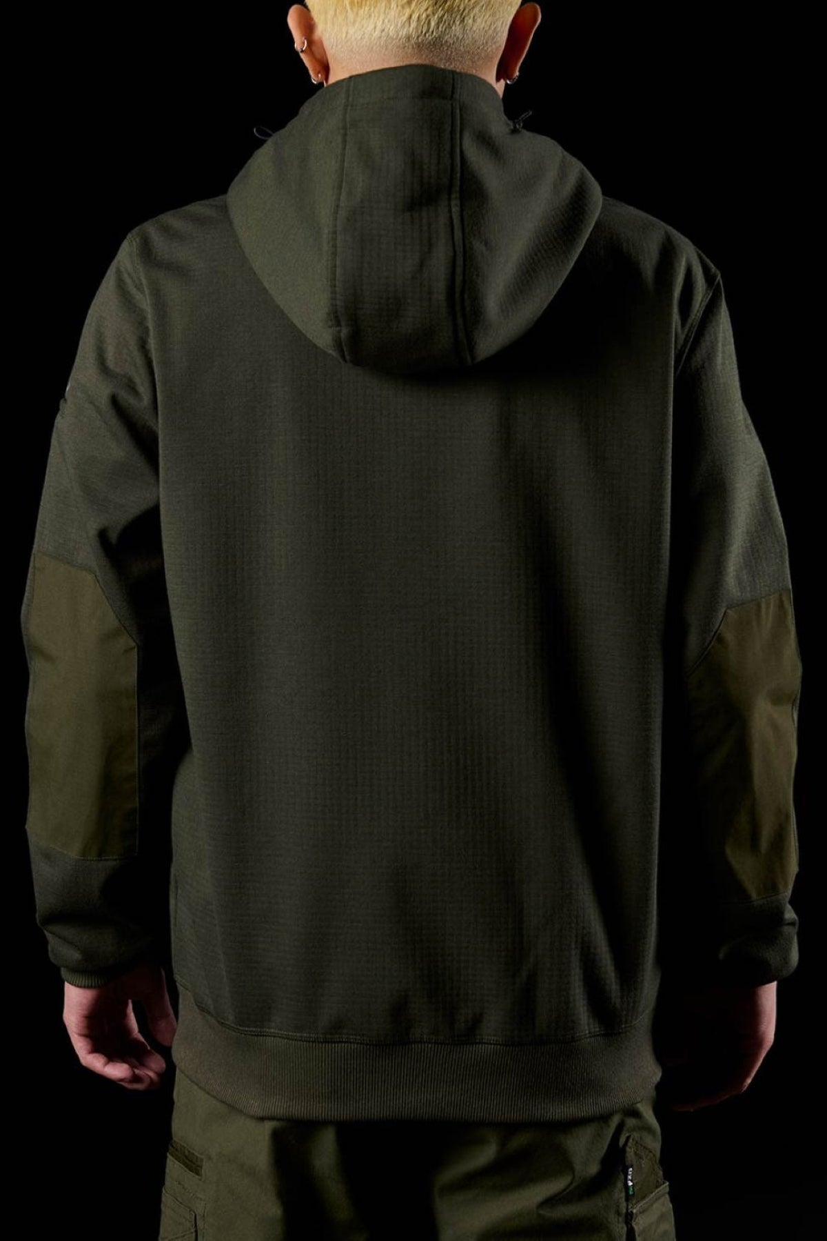 A person with short, light blond hair stands facing away against a black background, wearing a dark green FXD WF-3 Jacket and matching pants.