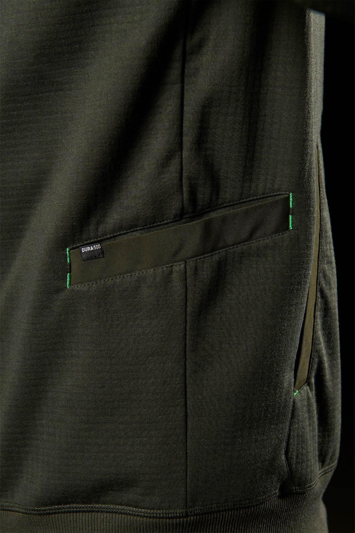 Close-up of a dark olive green FXD WF-3 Jacket pocket, featuring green stitching and a small black "Gunā 360" tag on the top edge, highlighting detailed workwear craftsmanship.