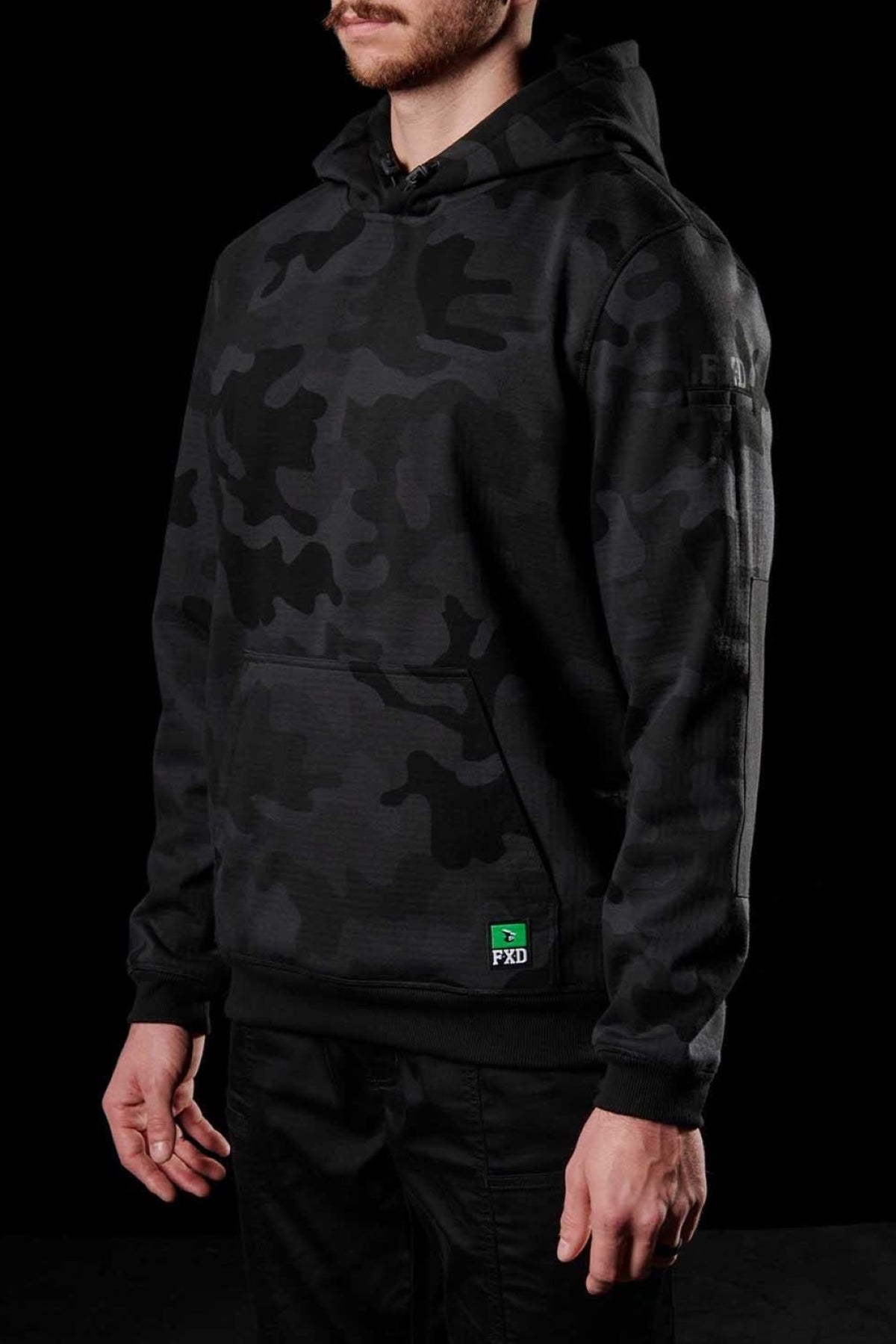 A man is positioned against a black backdrop, donning the FXD WF-1 Hoodie in a black and grey camouflage pattern. His hands rest casually by his sides. The hoodie features a green label near the hem with "FXD" printed on it. Only his torso and part of his arms are visible.