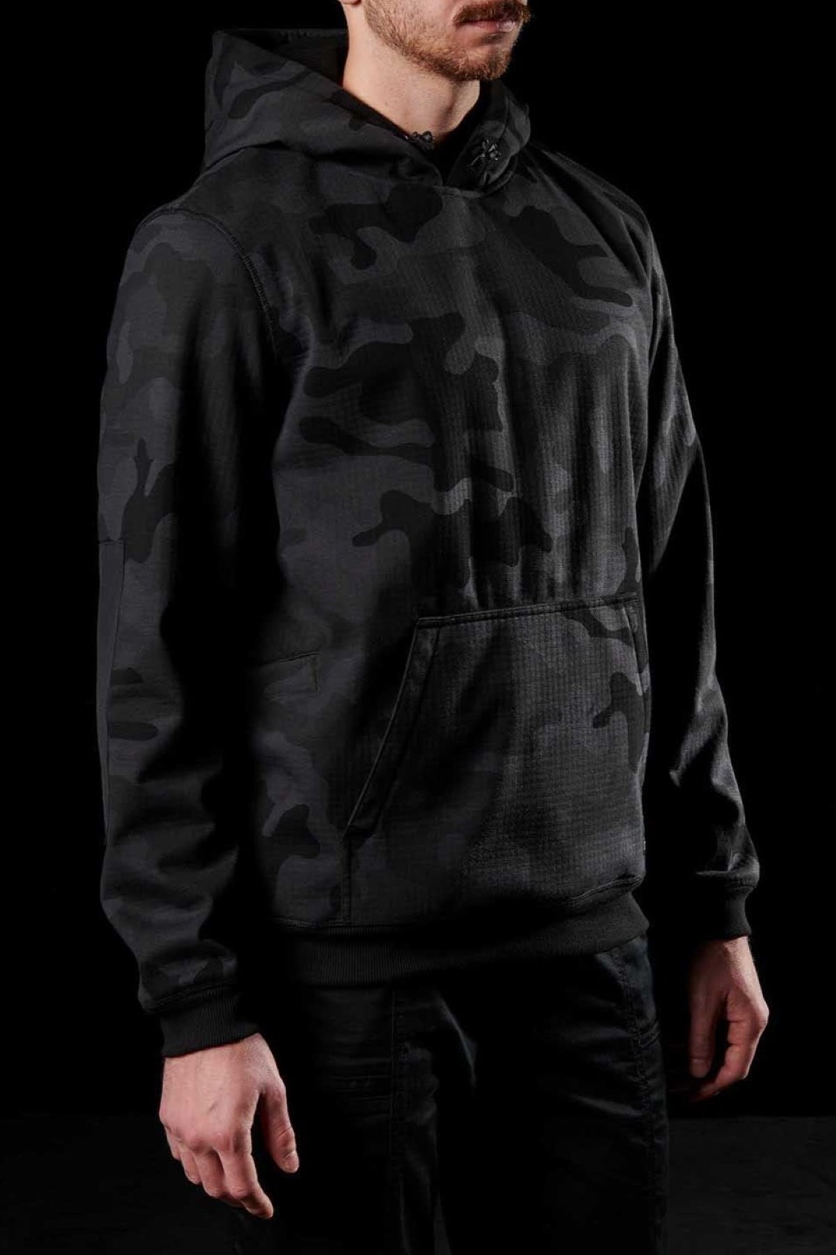 A person wearing an FXD WF-1 Hoodie stands against a black background. Only the lower part of their face is visible, and their hands are relaxed at their sides. The black windproof hoodie features a front pocket and a drawstring for adjusting the hood.