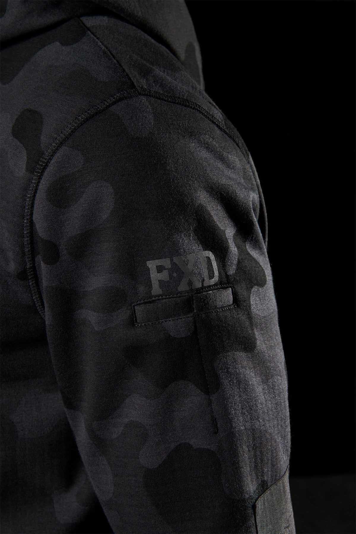 Close-up of a person's arm wearing an FXD WF-1 Hoodie in a black and gray camouflage pattern, with "FXD" printed on the sleeve, crafted from DURA500™ canvas. The background is completely black.