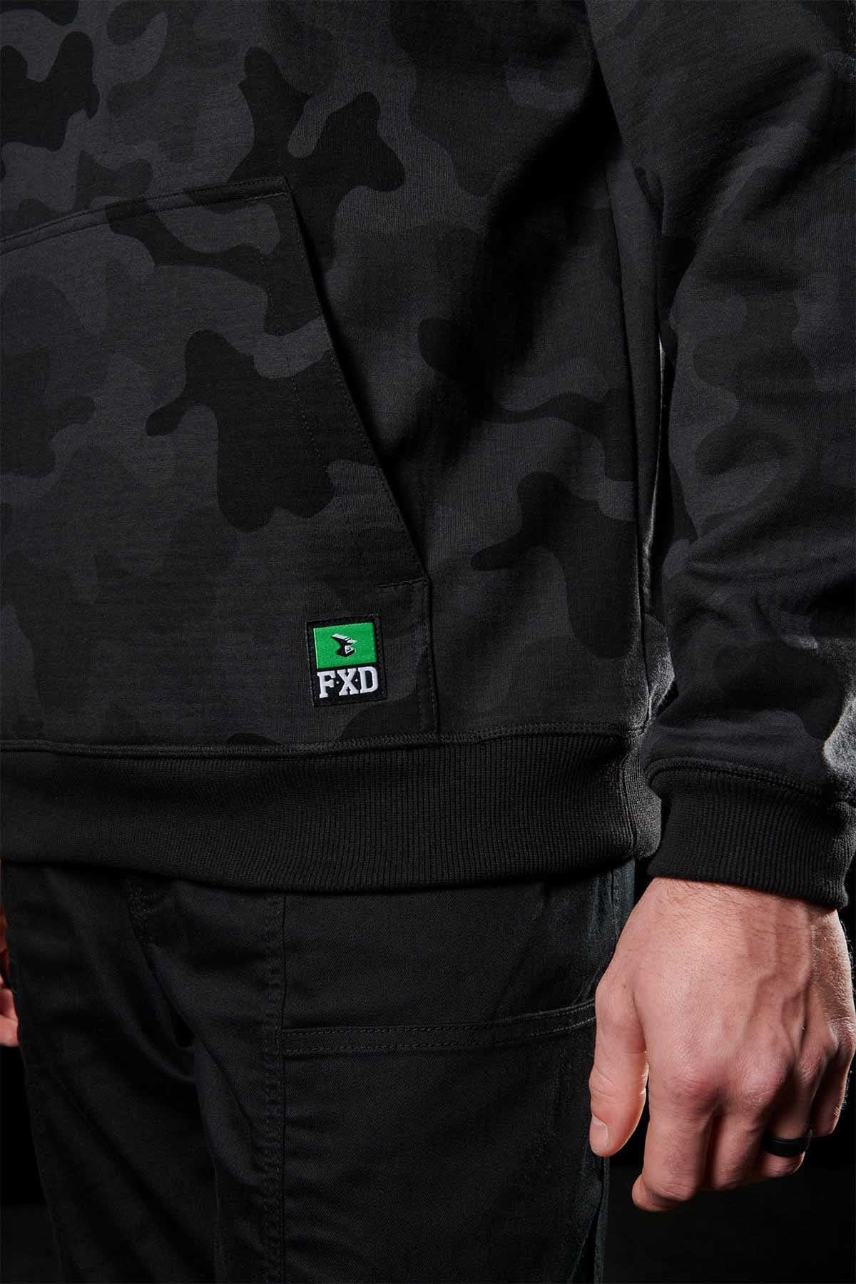A close-up image of a person wearing the FXD WF-1 Hoodie, which is black with a camo pattern and windproof material, paired with black pants. The hoodie showcases a small green patch with the letters "FXD" in white on the bottom left corner of the front pocket. The person is standing with their left hand visible.