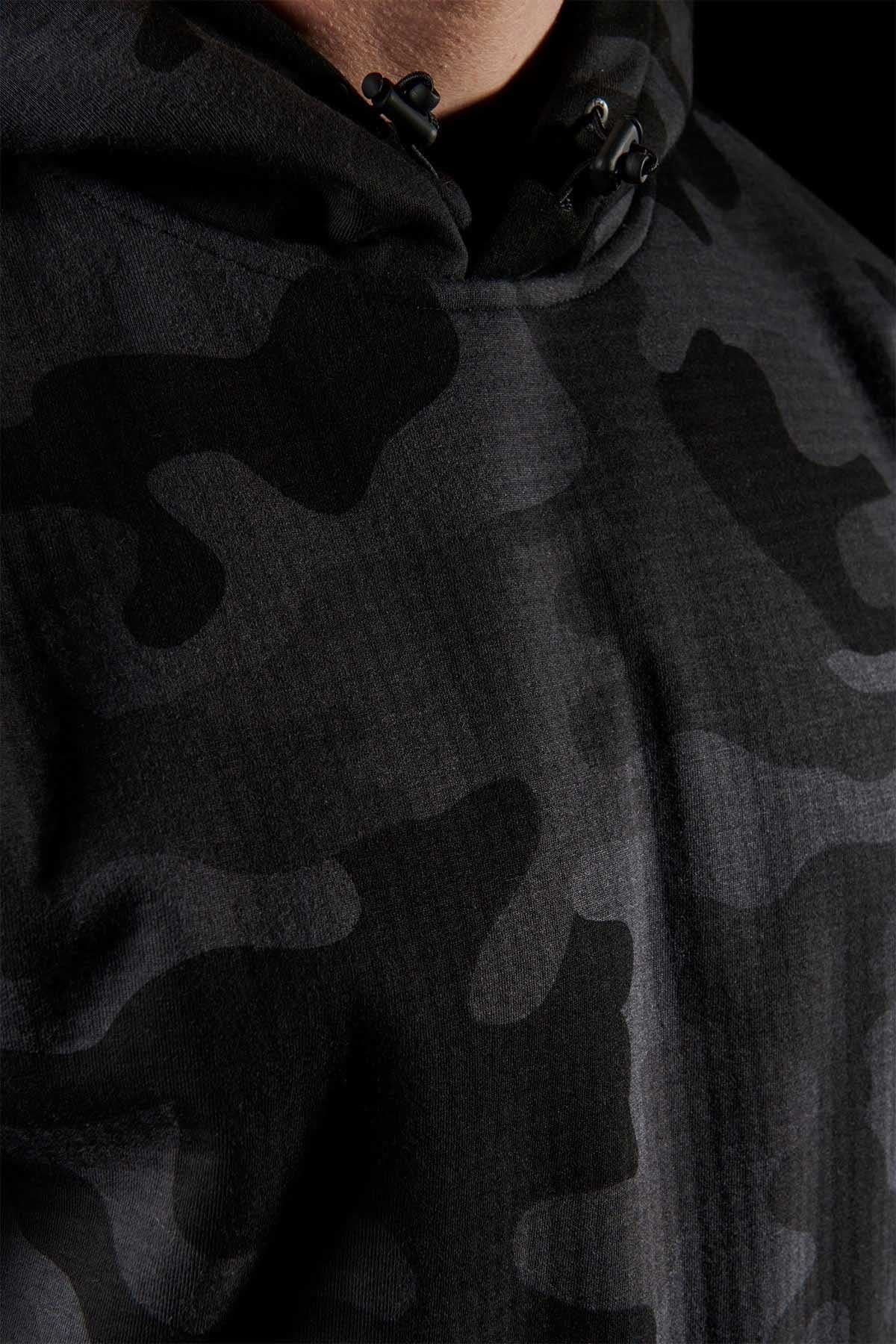 Close-up of a person wearing the FXD WF-1 Hoodie, a dark camouflage, windproof hoodie. The fabric showcases various shades of black and gray in irregular patterns. The hood is cinched at the neck with black drawstrings set against a matching black background.